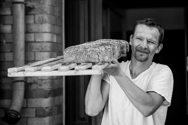 Ein Mann, der ein Brot auf einem großen Brett trägt. Klick führt zu Großansicht.