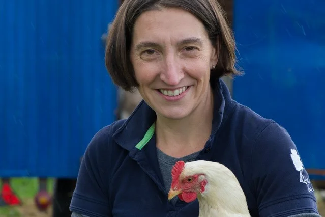Frau mit Huhn auf dem Arm lacht in Kamera. Klick führt zu Großansicht.