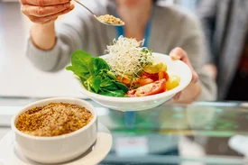 Eine Person hält einen Teller mit Salat an einer Ausgabetheke, während ein LöMel mit Topping darüber gehalten wird.