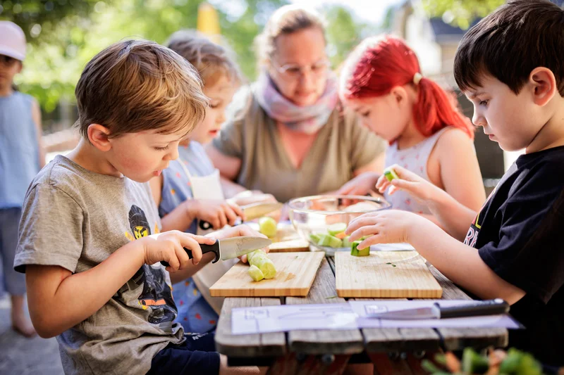 Kinder schneiden Gemüse am Tisch draußen. Klick führt zu Großansicht in Lightbox, Esc zum Schließen.