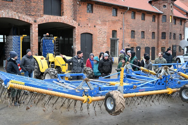 Junge Menschen stehen im Halbkreis um eine landwirtschaftliche Maschine und bekommen diese erklärt. Klick führt zu Großansicht.