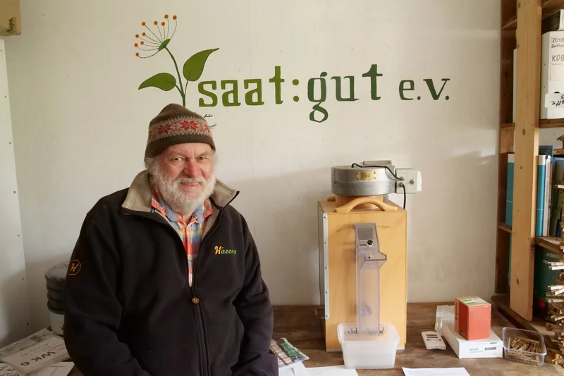 Mann vor einer Wand mit dem Vereinslogo "saat:gut e.V.". Klick führt zu Großansicht in neuem Fenster, schließen per ESC.