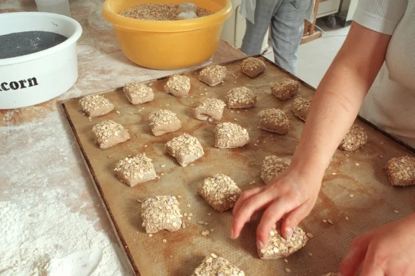 Zu sehen ist ein Backblech mit Körnerbrötchenteiglingen. Eine Hand platziert ein weiteres Brötchen auf dem Backblech. Klick führt zu Großansicht.