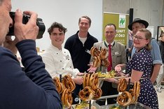Vier Männer und eine Frau stehen um eine Anrichte mit Brezen und Brotzeitbrettchen und lachen in die Kamera. Klick führt zu Großansicht.
