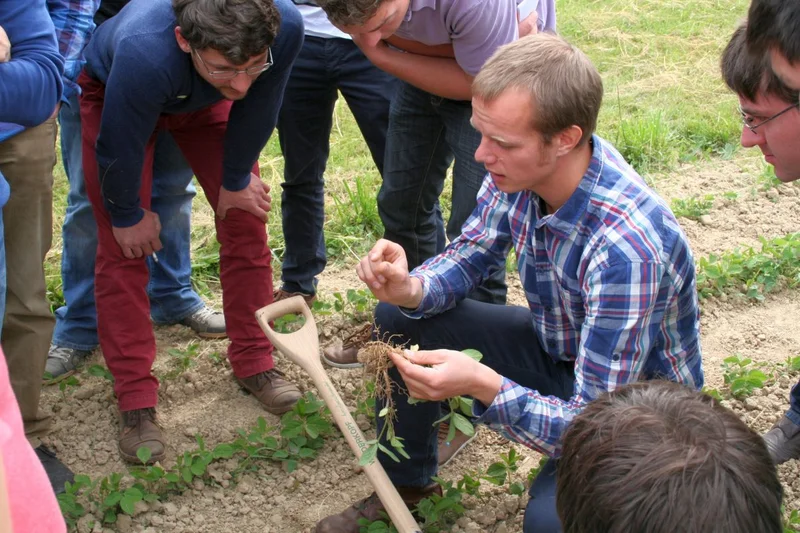 Ein Mann im Feld, wie er etwas erklärt, umringt von Menschen.