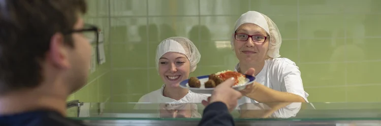 Zwei Frauen an der Essensausgabe in einer Mensa, die einen Teller über die Theke reichen.