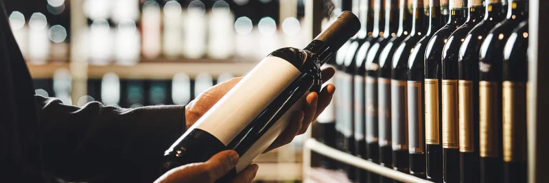 Mensch vor einem Weinregal mit einer Flasche Wein in der Hand.