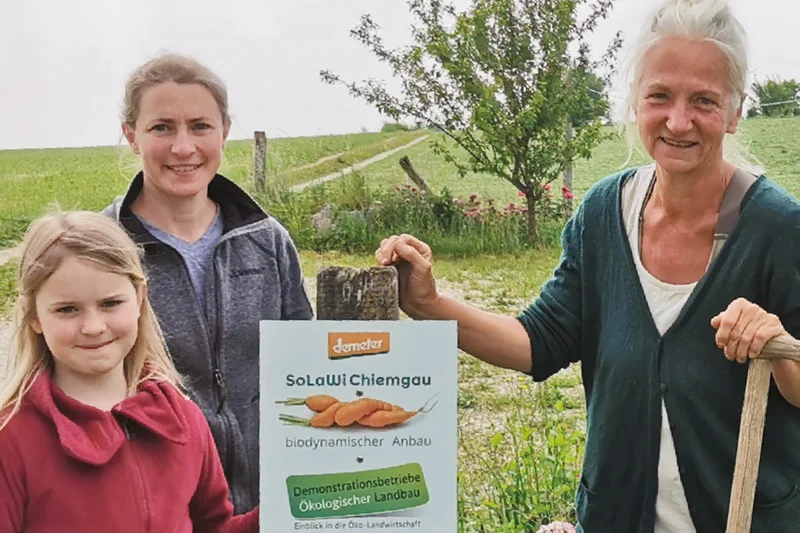 Betriebsleiterin Kristine Rühl rechts mit einer Frau und einem Mädchen am Zaun der SoLawi. Sie halten das Demo-Schild in der Hand