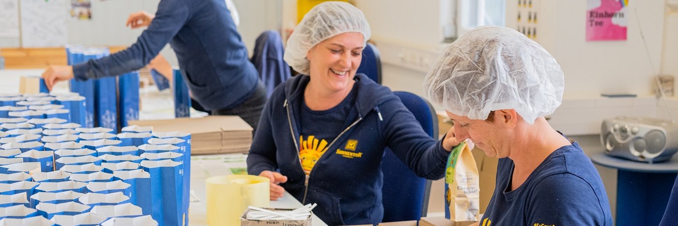 Drei Frauen nebeneinander mit Kopfhaube, die Kräuter in Tüten abpacken und dabei lachen.