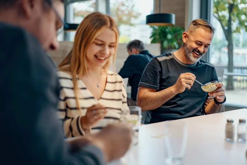 Mehrere Erwachsene sitzen an einem Tisch in einer Kantine und essen gemeinsam eine Mahlzeit. Klick führt zu Großansicht in einer Lightbox. Zum Schließen der Lightbox ESC drücken.