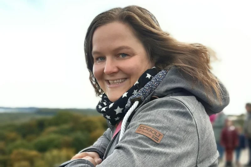 Frau stützt sich auf ein Geländer und lächelt in die Kamera.