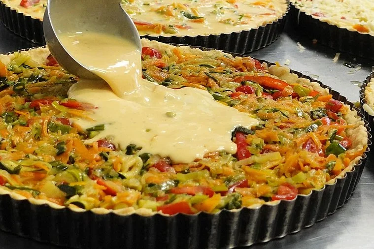 Eine Quiche in einer Backform auf die ein noch flüssiges Ei gegossen wird.