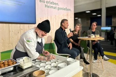 Zu sehen ist eine Frau, die Essen auf einer Messebühne zubereitet. Neben ihr sitzen ein Mann und zwei Frauen um einen Tisch. Der Mann hält ein Mikrofon in der Hand und erzählt etwas.