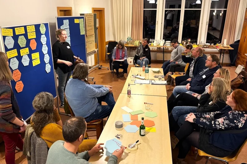Menschen, die um einen Tisch sitzen und diskutieren. Neben dem Tisch stehen verschiedene Pinnwände. Klick führt zu Großansicht.