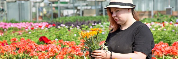 Frau steht inmitten von Blumen und hält eine Topfpflanze in den Händen.