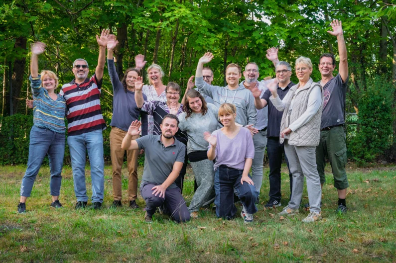 Gruppe von Menschen steht auf einer Wiese und winkt in die Kamera.