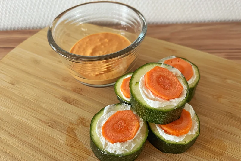 Zucchiniröllchen mit pikanter Ziegenkäsefüllung und Paprika-Dip auf einem Holzbrett serviert.