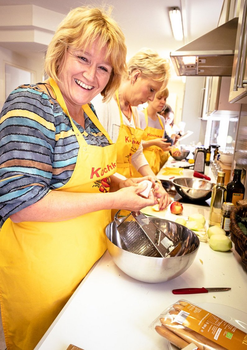Eine Frau in gelber Kochschürze und mit Käsereibe in der Hand lächelt in die Kamera.Hand
