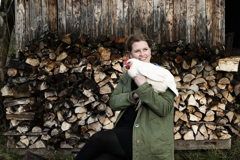 Eine Frau mit einem weißen Huhn auf dem Arm. Sie lacht in die Kamera. Kllick führt zu Großansicht.