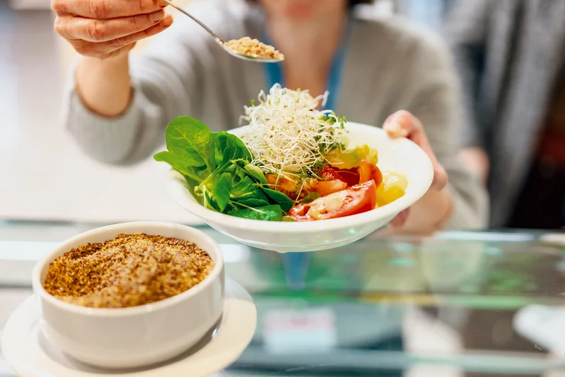 Eine Person hält einen Teller mit Salat an einer Ausgabetheke, während ein LöMel mit Topping darüber gehalten wird. Klick führt zu Großansicht in einer Lightbox. Zum Schließen der Lightbox ESC drücken.