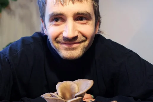 Mann lacht in Kamera. Vor ihm liegen Pilze auf dem Tisch. Klick führt zu Großansicht.