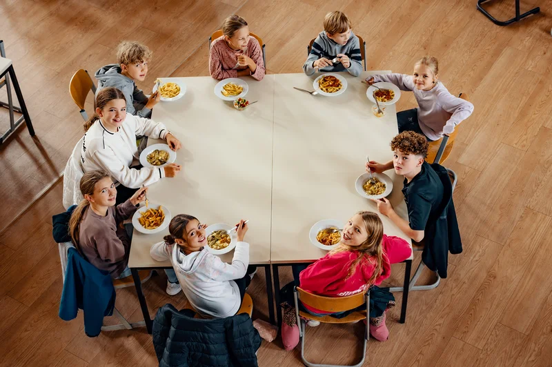 Neun Kinder sitzen an einem großen Tisch und essen gemeinsam; Blick von oben in einen Raum mit Holzfußboden.