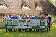 Menschen halten einen Banner mit der Aufschrift "Öko-Feldtage 2027 – Bauckhof Amelinghausen". Sie stehen vor einem Haus mit Strohdach und lachen in die Kamera. Klick führt zu Großansicht.