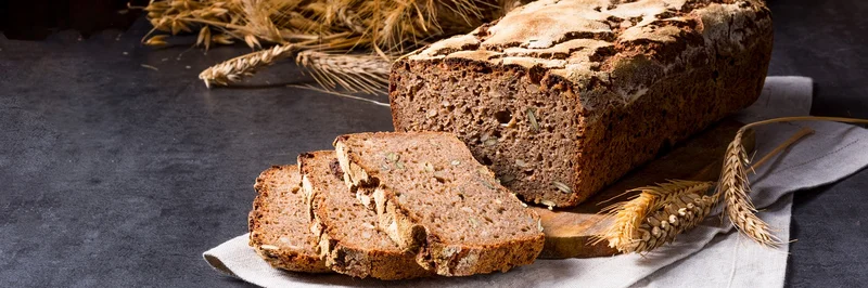 Ein aufgeschnittenes Brot. Die Scheiben liegen vor dem Brot. Im Hintergrund sind Getreideähren zu sehen.