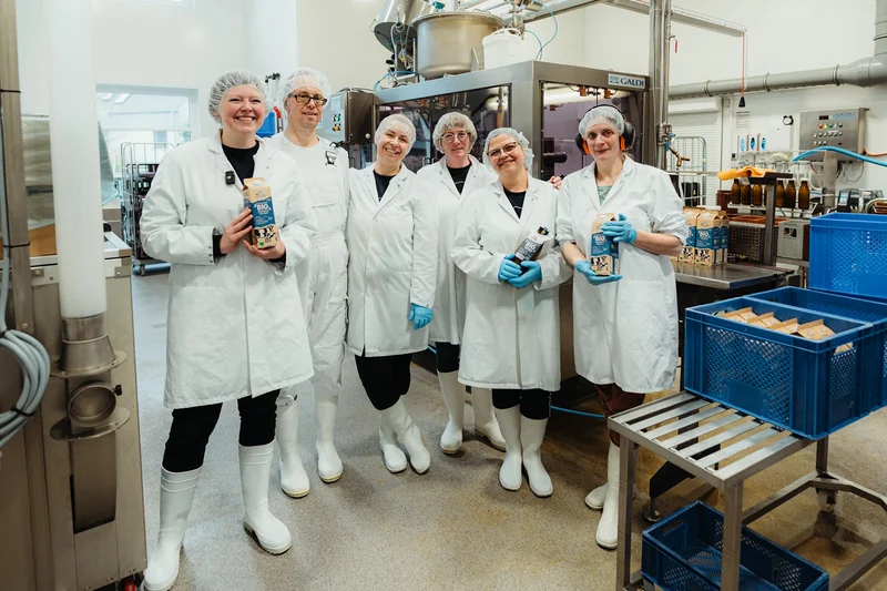 Mehrere Menschen mit Laborkitteln und Milchprodukten stehend in einer Molkerei. Klick führt zu Großansicht.