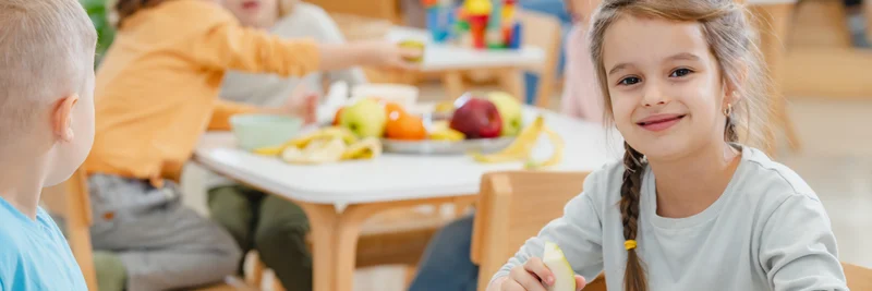 Kinder in einer Kita an verschiedenen Tischen, auf denen Obst in einem großen Teller steht. Ein Mädchen hat eine Birne in der Hand und lacht in die Kamera. Ein Junge mit einer Birne in der Hand schaut nach hinten zu den anderen Kindern.