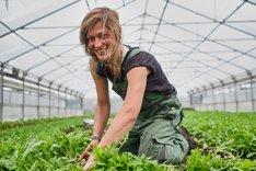 Frau in einem Gewächshaus auf dem Boden bei der Pflanzarbeit. Sie schaut freundlich in die Kamera. Klick führt zu Großansicht.