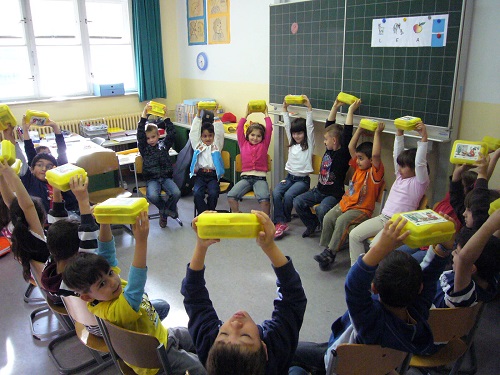Kinder in der Schulklasse halten gelbe Brotdosen. Klick führt zu Großansicht im neuen Fenster.
