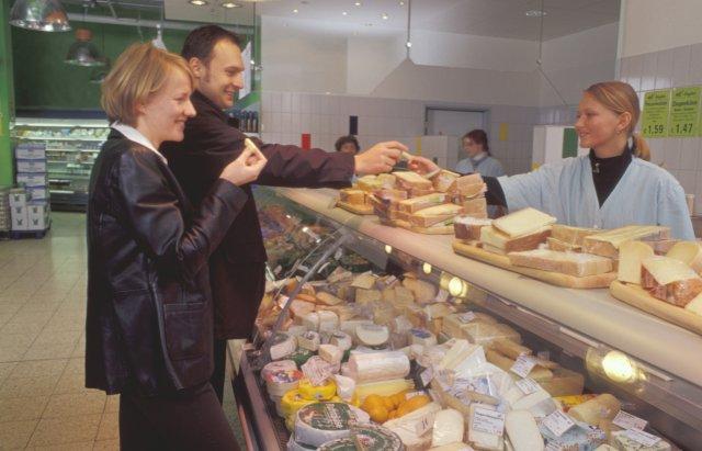 Eine Verkäuferin reicht einer Kundin und einem Kunden an einer Käsetheke Käsestückchen zur Verkostung. Klick führt zu Großansicht im neuen Fenster.